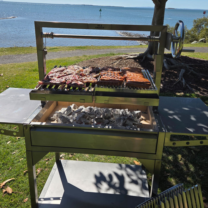 Front view of an OLA Grills Parrilla BBQ cooking pork chops and sausages by the waterfront